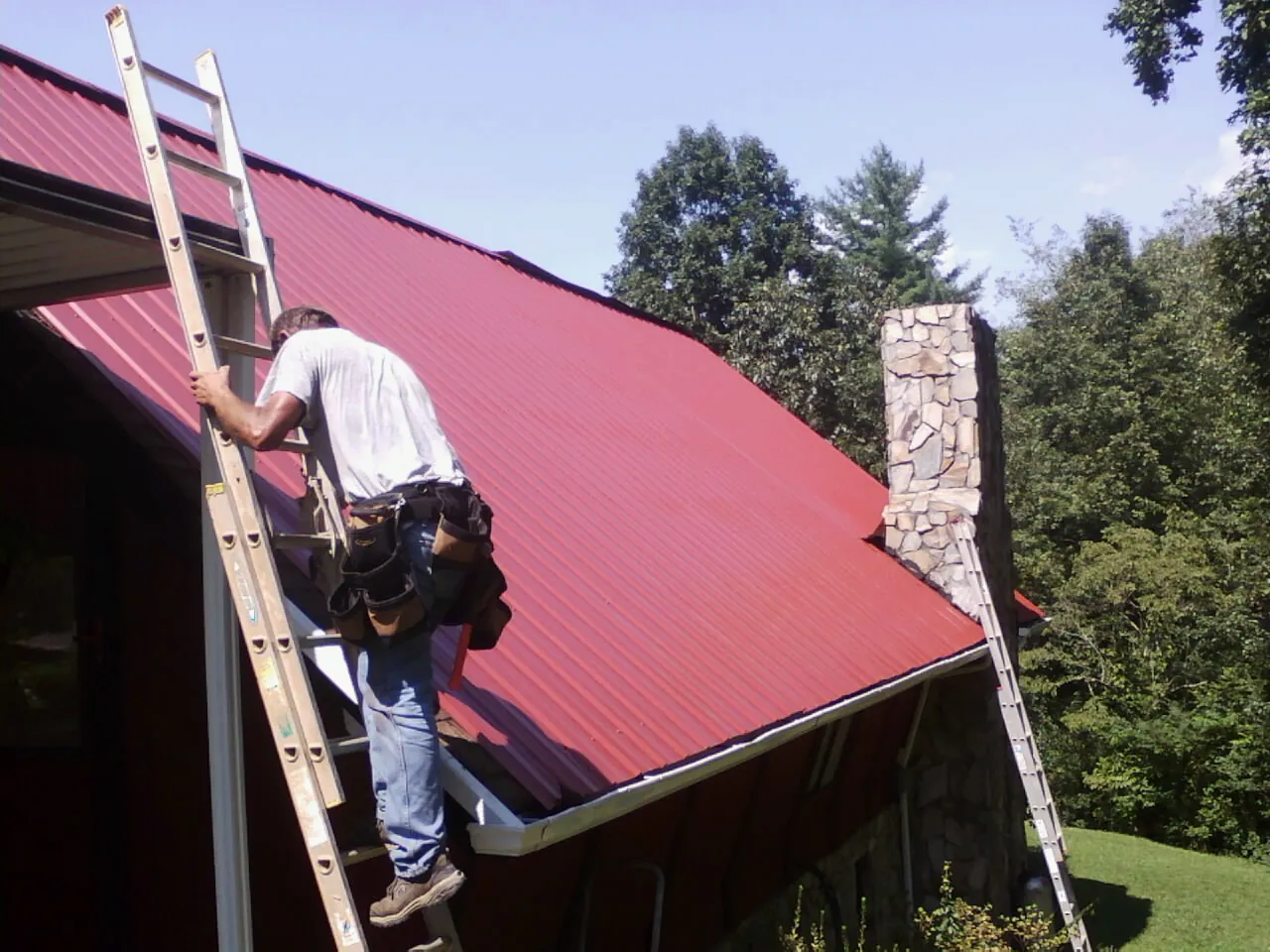 Roofer on a ladder beside a red metal roof and stone chimney in Little Rock